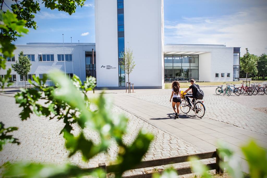 Persons walking and biking outside a modern white building on a sunny day.